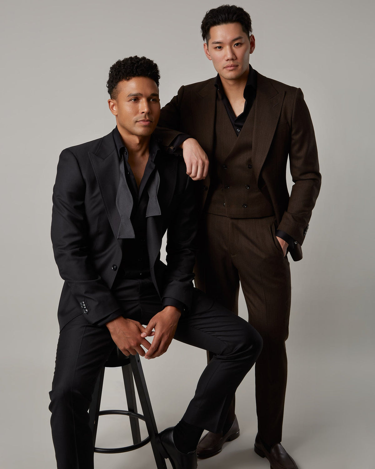 Black suit worn seated beside a brown three-piece with double-breasted waistcoat, from the ELEVATE shoot