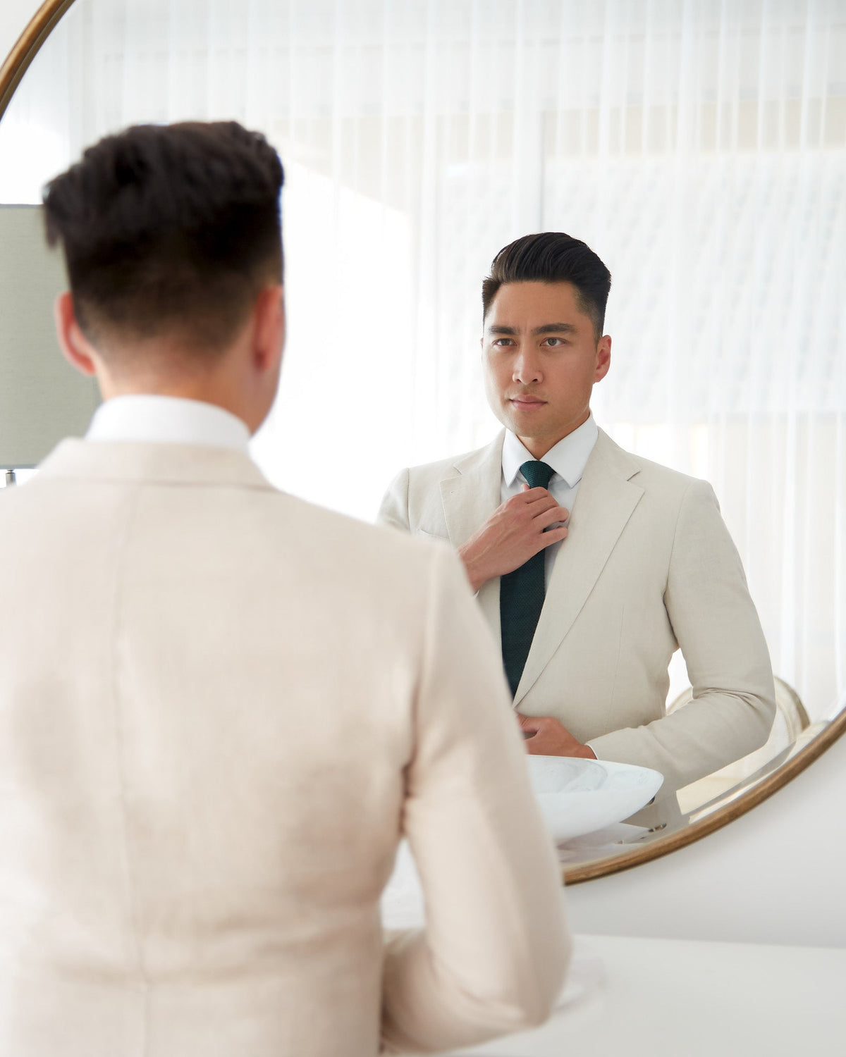 Man in a cream made to measure suit straightening a dark green tie in a round mirror