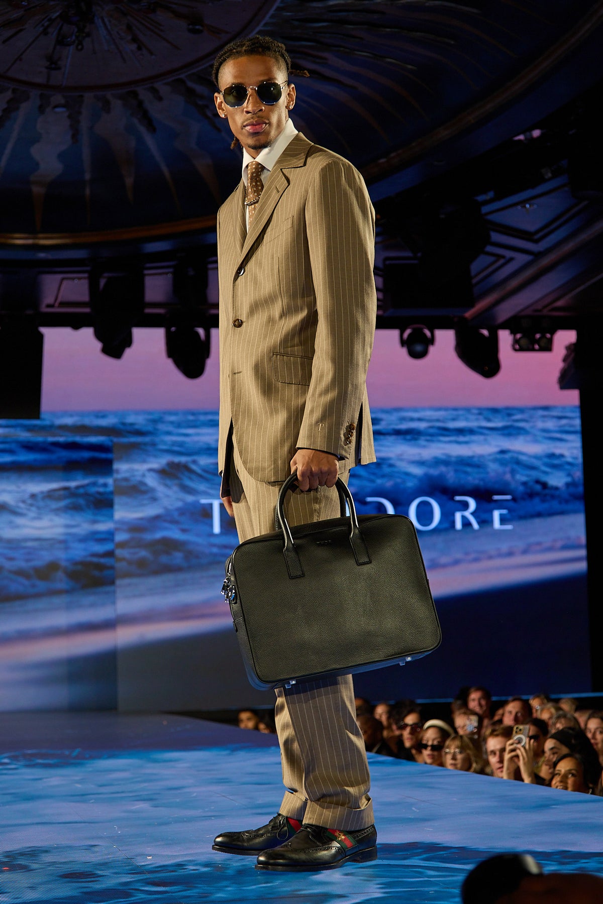 Beige Pinstripe Suit, a model walks the runway past an ocean-themed backdrop with a black bag