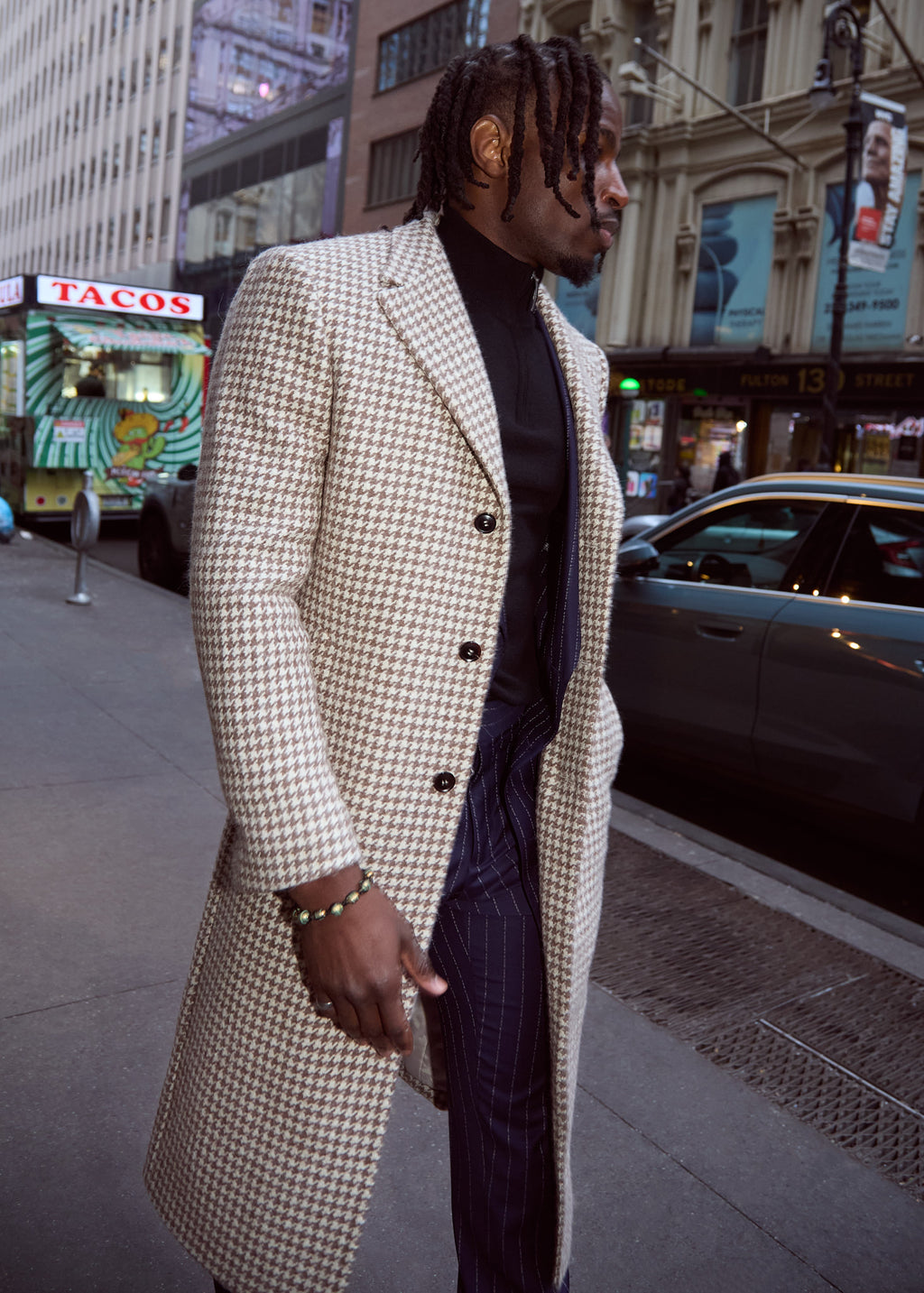The No Filter Navy Pinstripe Suit trousers beneath a houndstooth overcoat, model on a city street