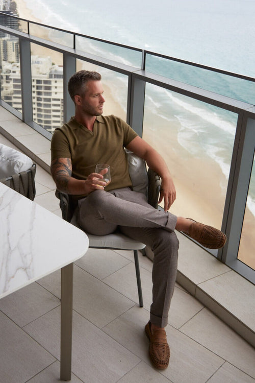3J Whisky Shot Polo Olive worn by a seated model on an ocean-view balcony