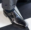 The Friar Black Leather Double Monk worn on a model's foot on a checked floor