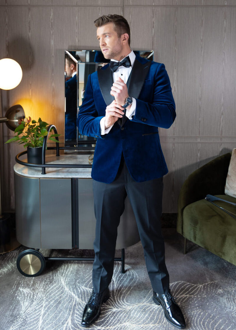 Velvet Noir navy tuxedo worn on a model adjusting his cufflink by a bar cart
