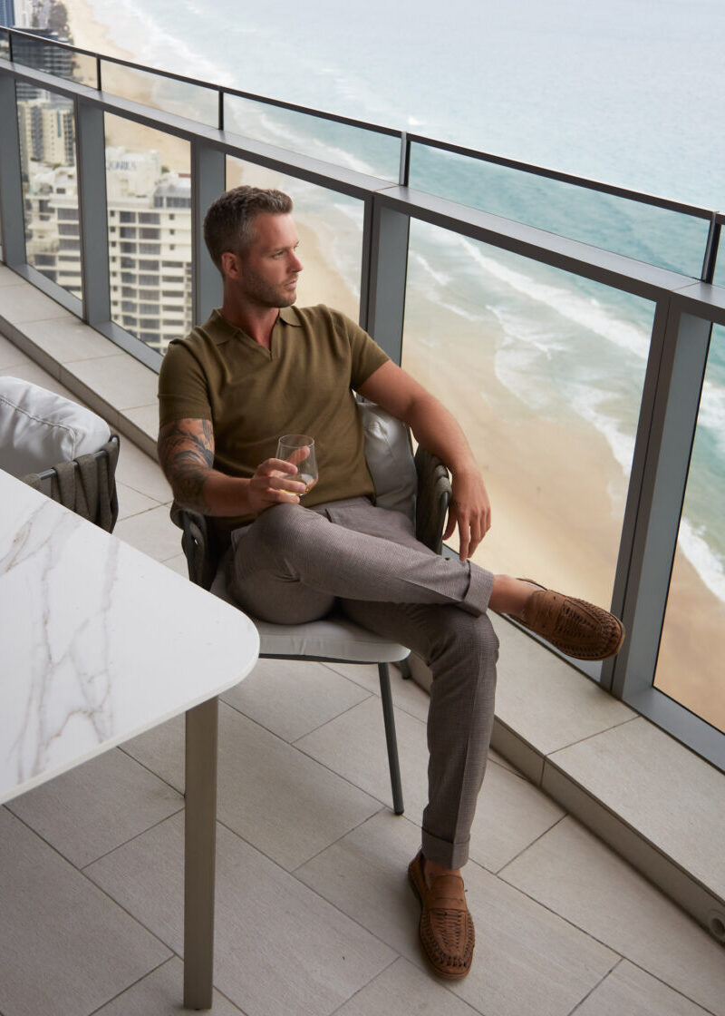3J Whisky Shot Polo Olive worn by a seated model on an ocean-view balcony