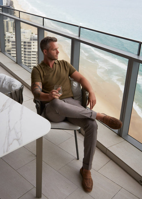 3J Whisky Shot Polo Olive worn by a seated model on an ocean-view balcony