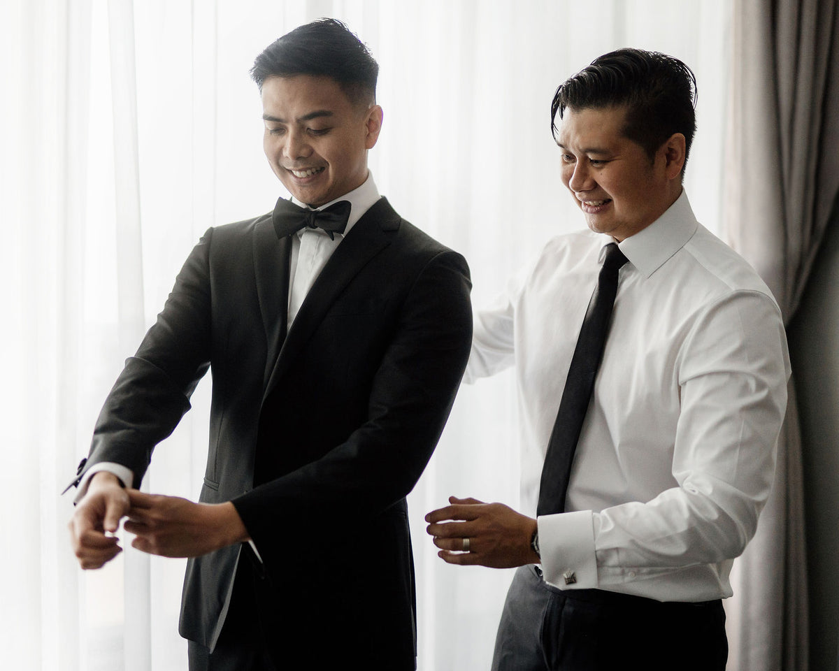 A groom in a black tuxedo fastening his cuffs beside a groomsman